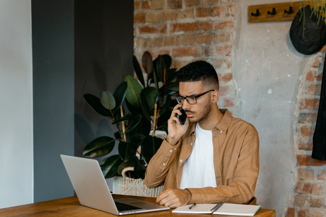Home Young man in glasses working on a laptop and phone in a modern office setting.