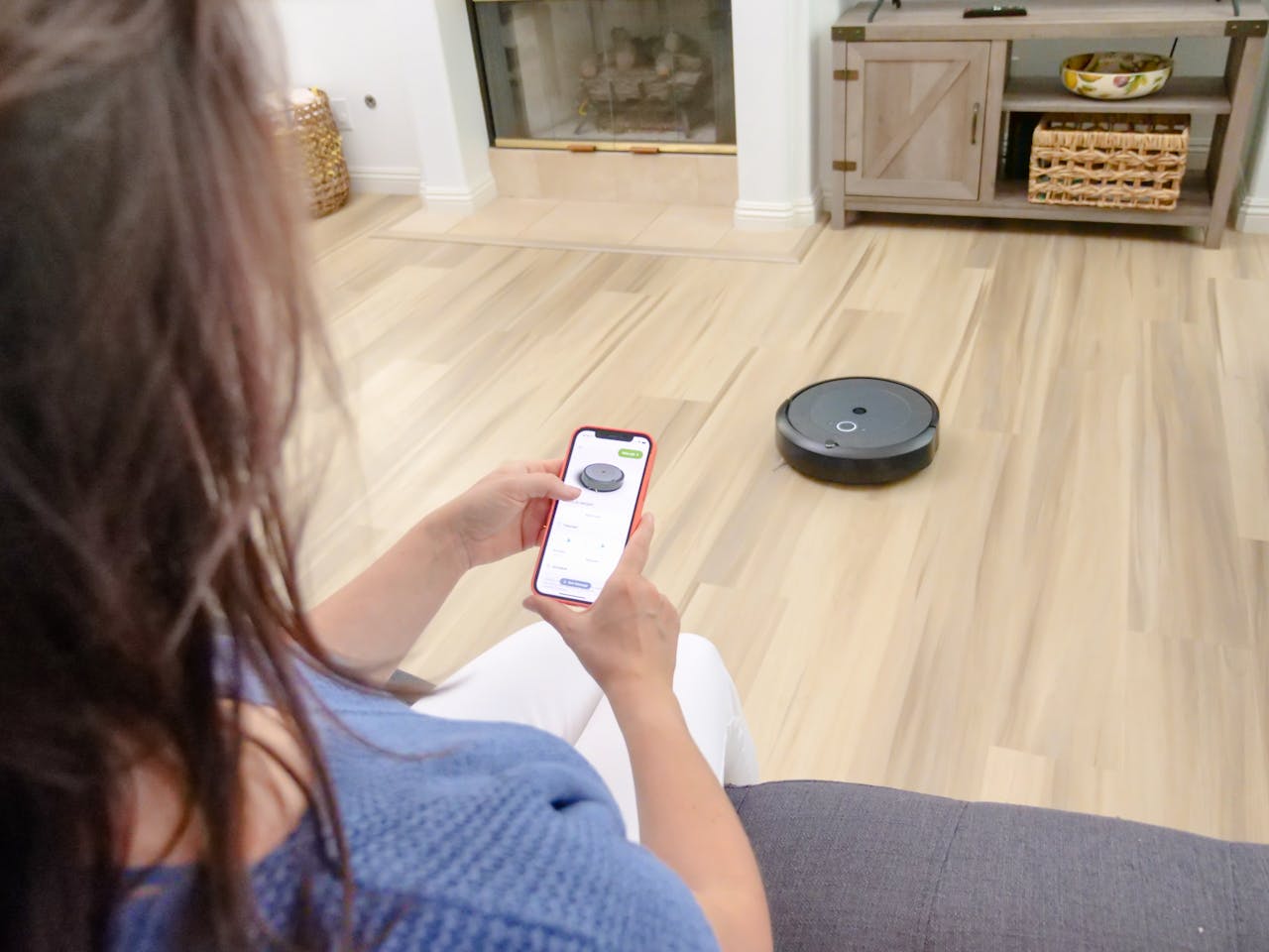The Art of Drawing Readers In: Your attractive post title goes here Overhead view of a woman controlling a robot vacuum using a smartphone app on a wooden floor.